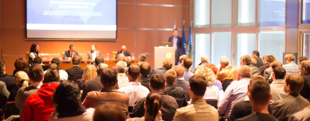 Crowd of people sitting and watching a man behind a podium delivering speaking with a presentation displayed on a screen